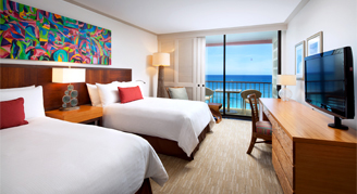 A view of a room with two beds, a desk and a T.V. with a view of the ocean in the background.