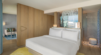 The image shows a king-size bed with white linens, wood-paneled sliding partitions, and a partial view into the modern bathroom area.