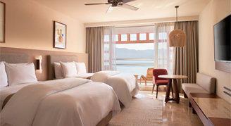 Interior view of the Partial-Ocean-View Junior Suite with two double beds overlooking the trees and ocean.