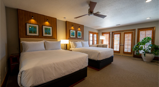 A spacious hotel room featuring two queen beds with crisp white linens, warm wood headboard panels.