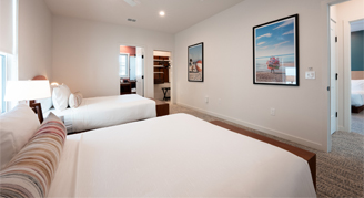 The image shows a hotel bedroom with two white-sheeted beds, light walls, and framed artwork hanging on the wall.
