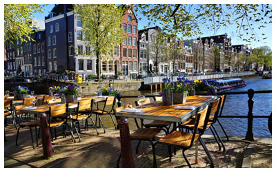 Restaurant tables lining the beautiful canals of Amsterdam under blue skies.